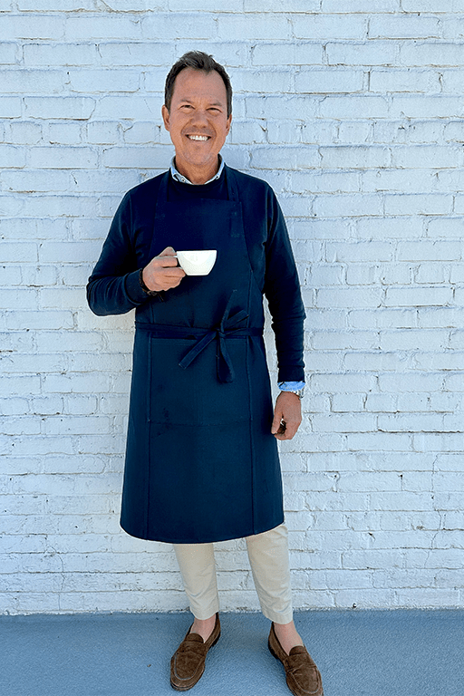 An image of a man wearing a denim apron standing in front of a brick wall drinking a cup of coffee.